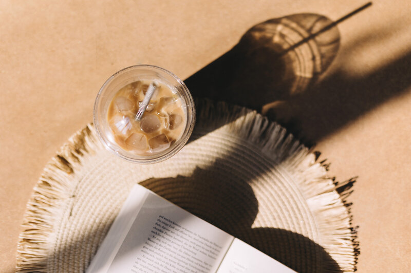 Flat lay of an iced coffee with a straw on a woven coaster beside an open book on a round table, casting natural shadows. Represents calm and clarity—mirroring the ease and support of two weeks of Slack-based implementation guidance after your business audit.