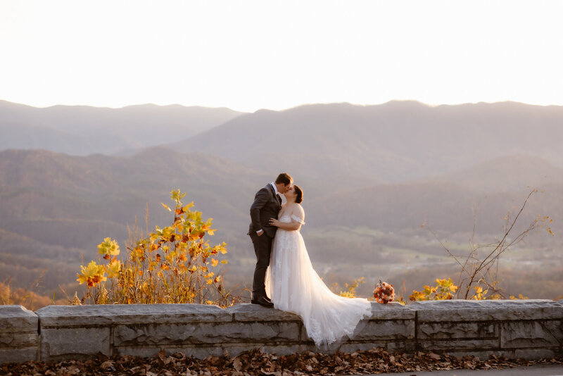 couples eloping to Gatlinburg in the Foothills with bride and groom embracing and kissing on a stone wall with the Smoky Mountains behind them photographed by photographers in gatlinburg tn