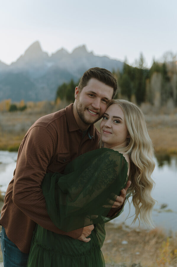 Romantic couples portraits in the Grand Tetons. With his hand behind his fiancé's back and touching foreheads, a man and a woman smile at the camera.