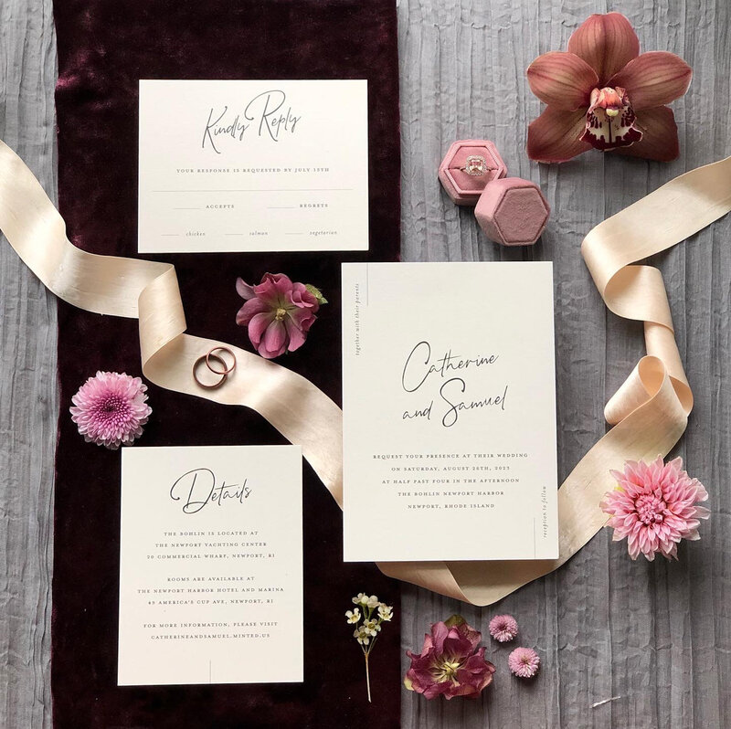 A selection of invitations, enclosure cards, and engagement ring in a velvet ring box, and wedding flowers featuring orchids, hellebores, wax flower and chrysanthemums rest on silk ribbon, burgundy velvet, and gray crinkle linens