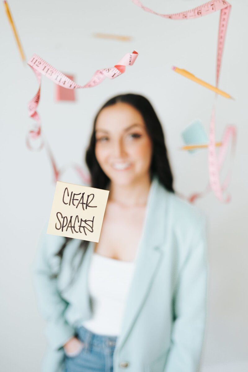 Woman in pastel blue blazer smiles at the camera with her hand in her pockets as pencils, post its and measuring tapes are suspended above her. A yellow sticky note reads "Clear Spaces"