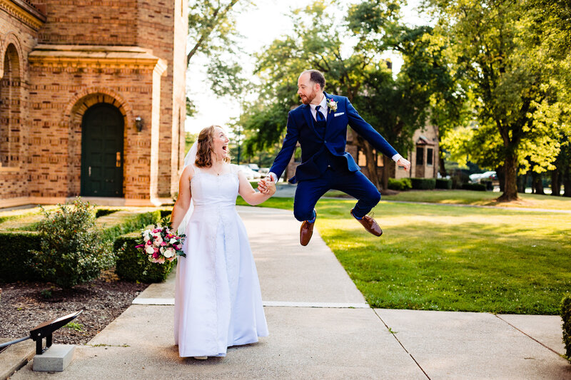 A wedding couple walking hand in hand as the groom jumps in the air as the bride gasp in laughter at Nazareth Hall in Toledo Ohio