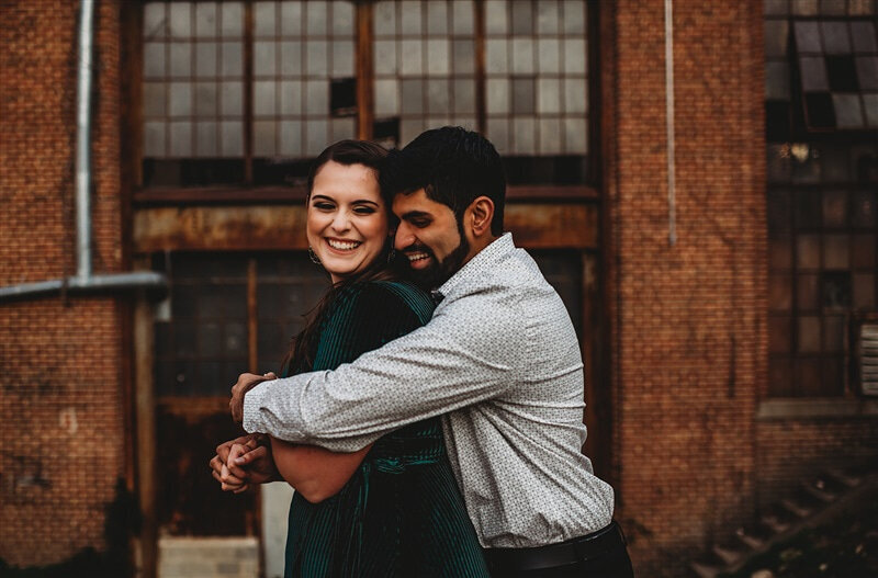 Baltimore photographer captures engagement photos with man standing behind a woman and a green dress and hugging her around the shoulders as they both laugh