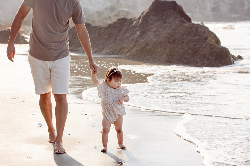 Father holding toddler daughter's hand walking along the shore of Avila Beach, CA with Fresno, CA family photographer angelia jansen