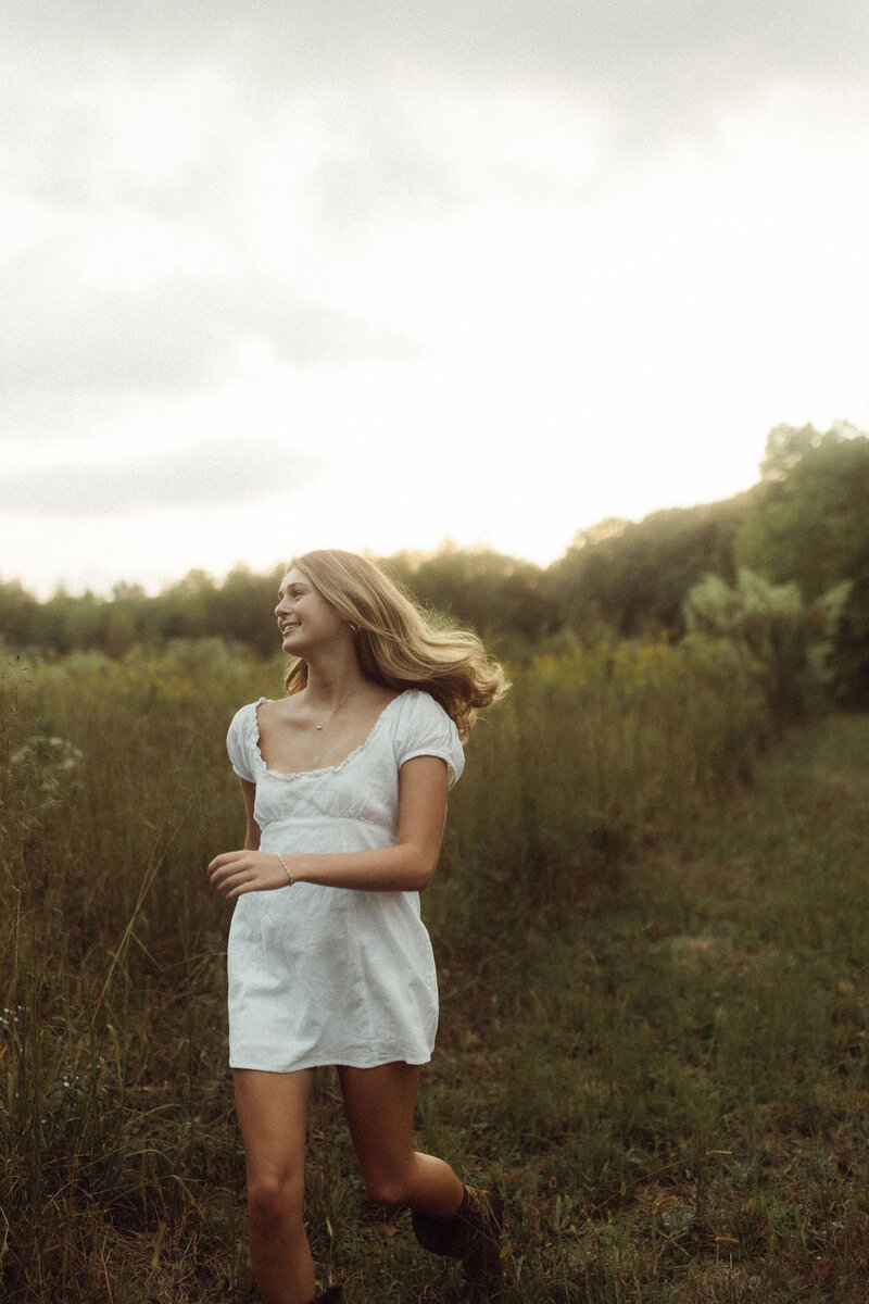A senior girl in a white dress runs through a lush green field.