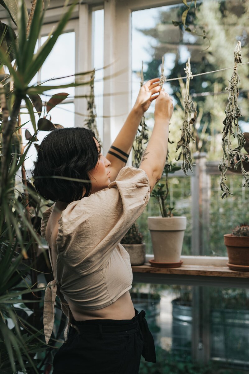 Woman stands in lush, greenhouse wearing a tan crop top and black pants while reaching up to hang herbs to dry on a line upside down.