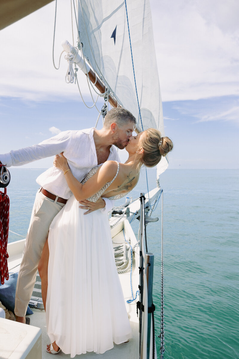 bride and groom embracing while standing on the beach with the ocean and a sailboat in the background