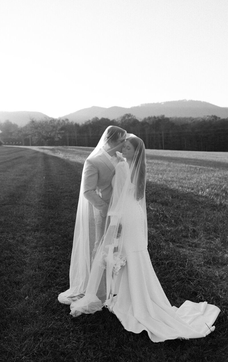 Newlyweds snuggle and kiss under the veil while standing in a pasture at sunset