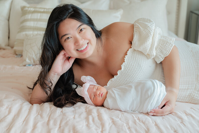 Mom smiles while lying on bed with baby girl in white swaddle.