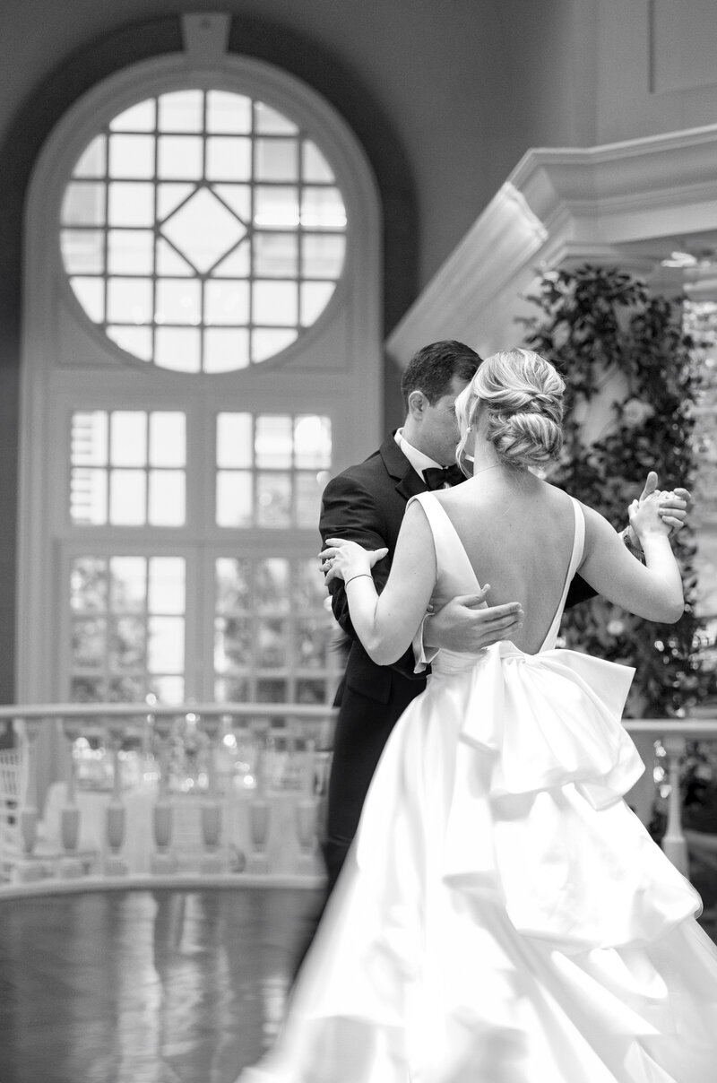 A couple dances elegantly in a beautiful indoor setting. The bride wears a white gown with a large bow, while the groom is in a suit. They are near a large, arched window with natural light streaming in.