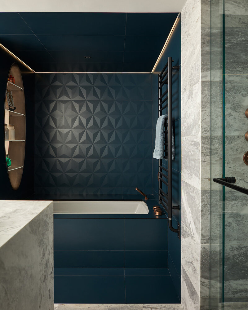 A luxury bathroom featuring built-in cove lighting, a tub surrounded by deep blue 3D tiles, and a marble countertop vanity and shower area