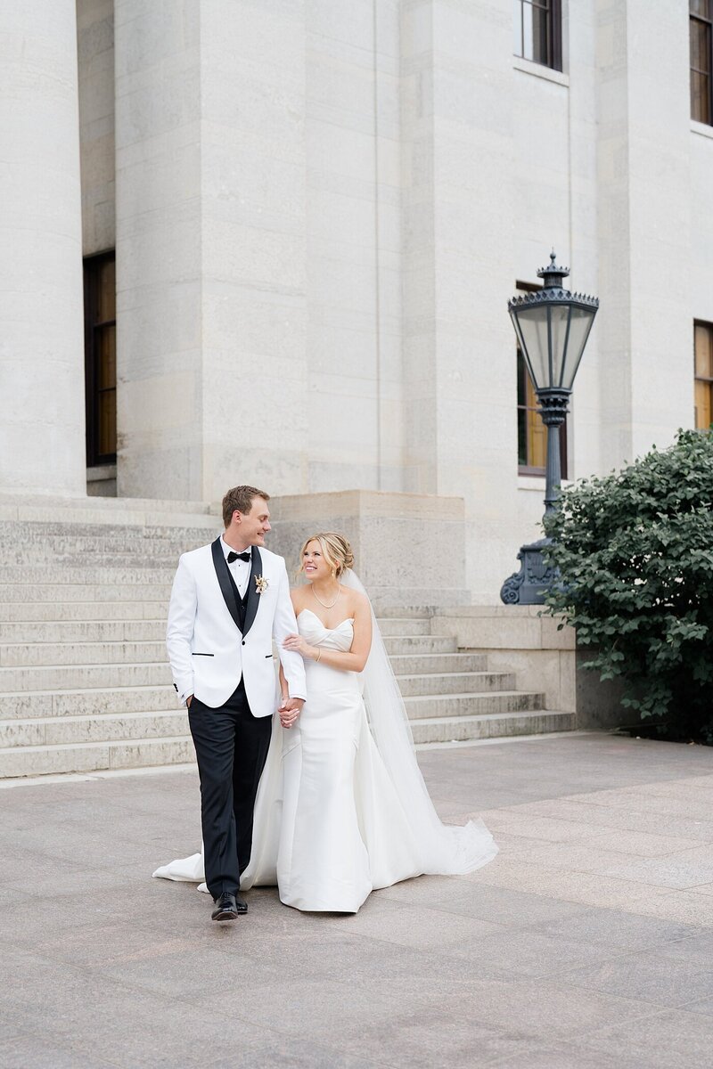 wedding photos by Columbus wedding photographer with bride holding onto her grooms arm and smiling at him as they walk together at the Ohio state Capital building