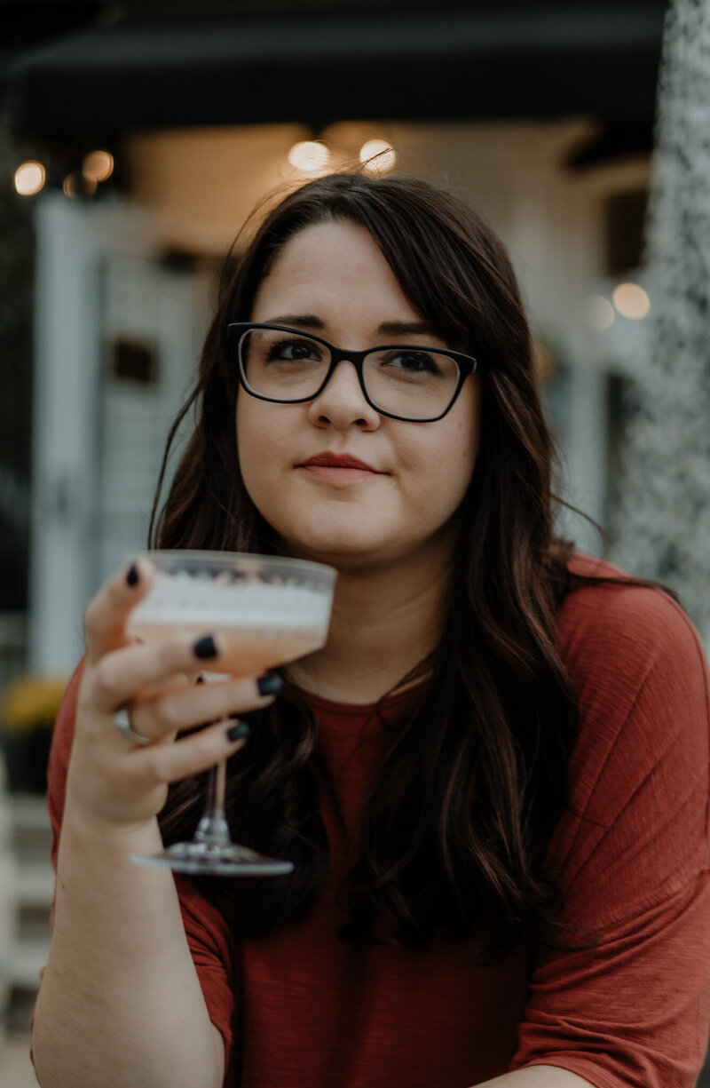 woman with glasses in terracotta shirt holding a martini glass looking off into the distance