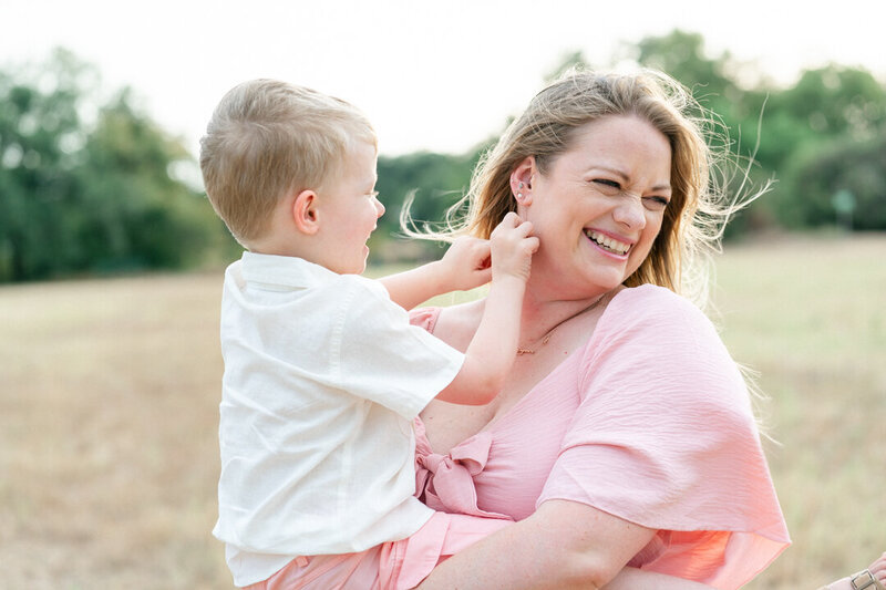 a mother dressed in a pink dress holds her toddler boy while he tickles her and she laughs.