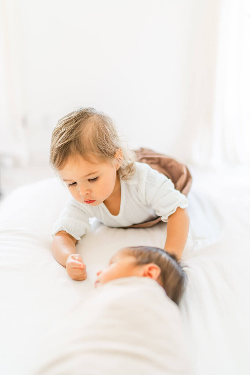 a toddler girl lays on a white bed and goes up to her newborn brother touching the tip of his nose.
