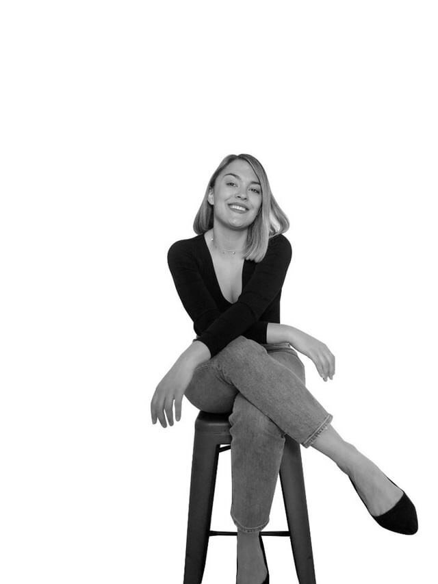 A woman with short blonde hair sits on a stool with her legs and arms crossed.