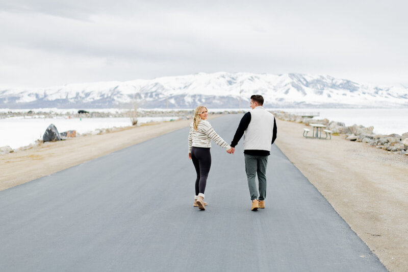 couple walking away from the camera along a road on a lake jetty, girl is looking back and smiling
