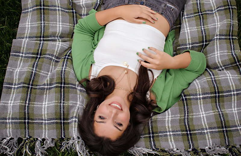 High school senior in a green cardigan, white crop top, and gray jeans lying on a plaid blanket and looking up at the camera with a relaxed expression, captured in a spring portrait session in South Charlotte NC.