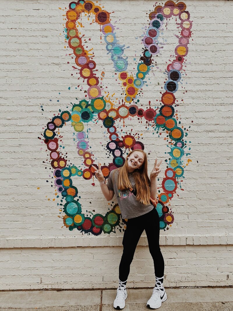 Teen girl brown hair in black pants in front of peace sign on wall 