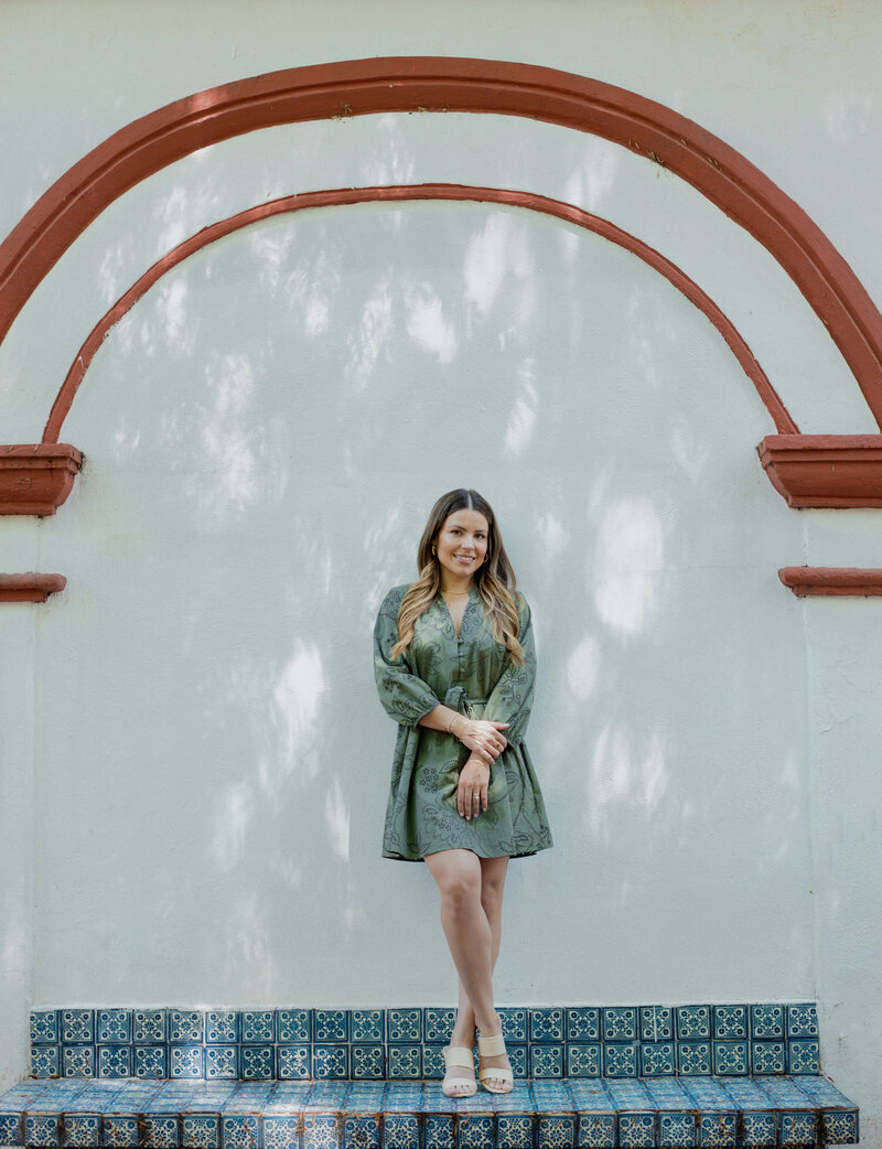 Natalie Bernacchi standing under a red archway in a green dress, smiling and grounded on decorative blue tile steps