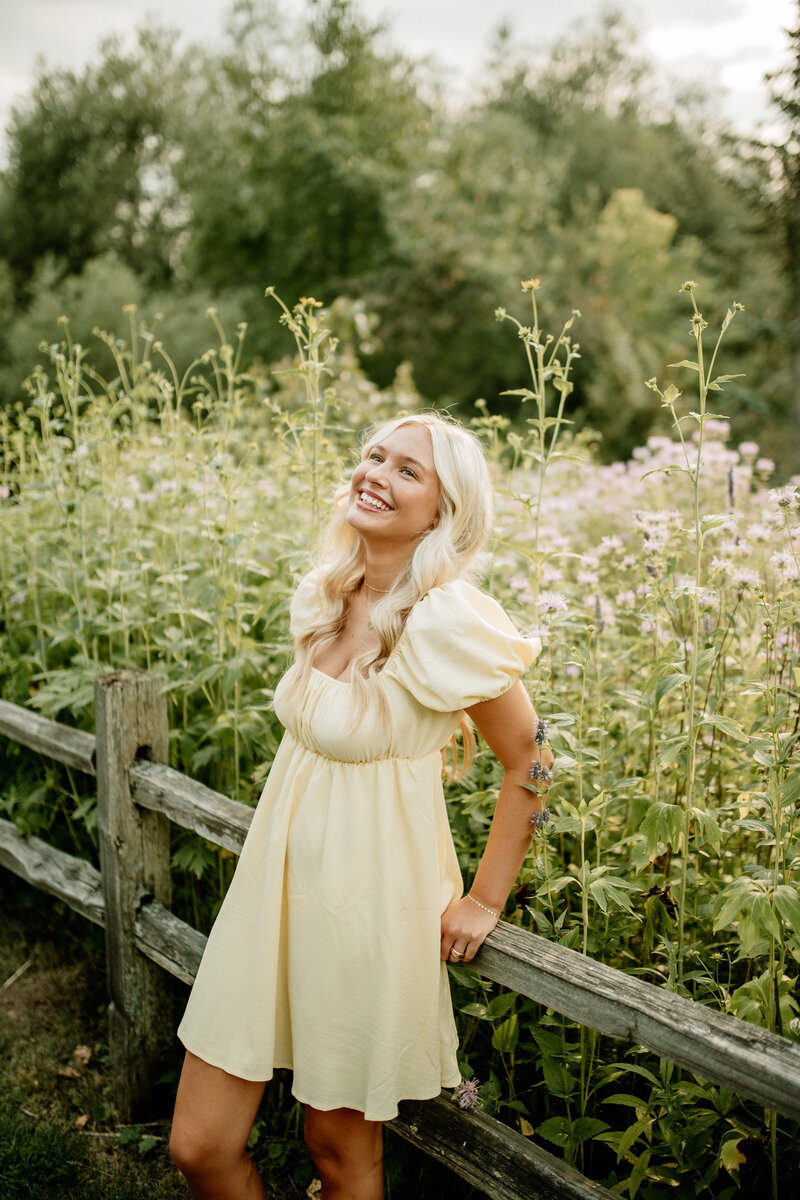 A high school senior leans against a wooden fence among wildflowers during summer senior portraits.