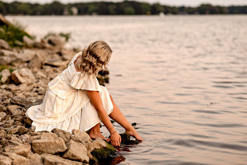 Senior girl in a white dress sitting at the edge of a lake dipping her toes in the water.