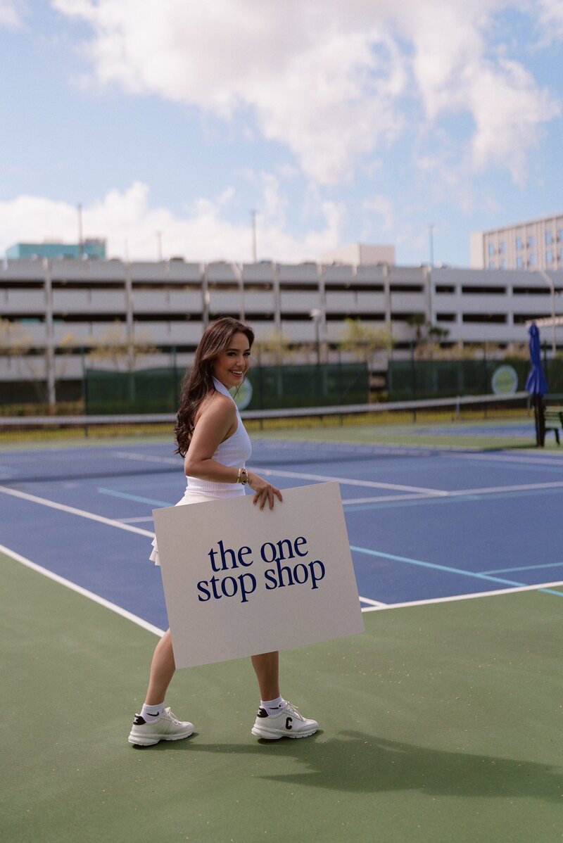 Woman walking on tennis court holding sign saying the one stop shop