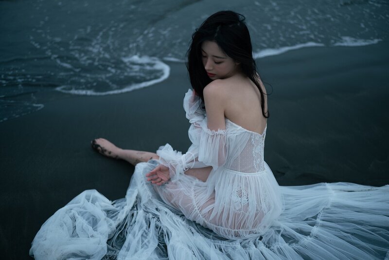 A woman wearing a delicate sheer couture dress shot on the beach in dark moody tones.