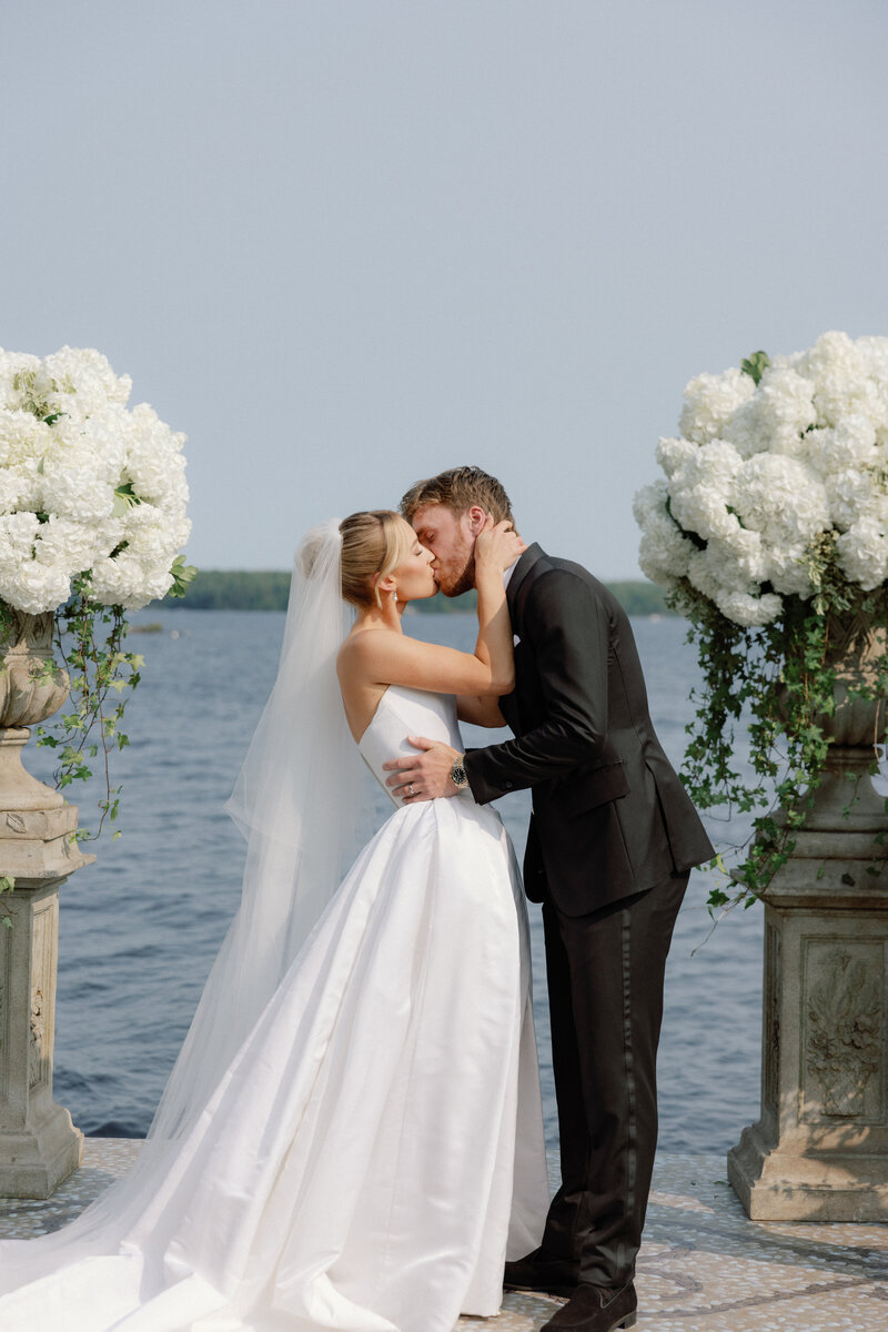 A bride pours a bottle of champagne in the top glass of a champagne tower at an outdoor venue