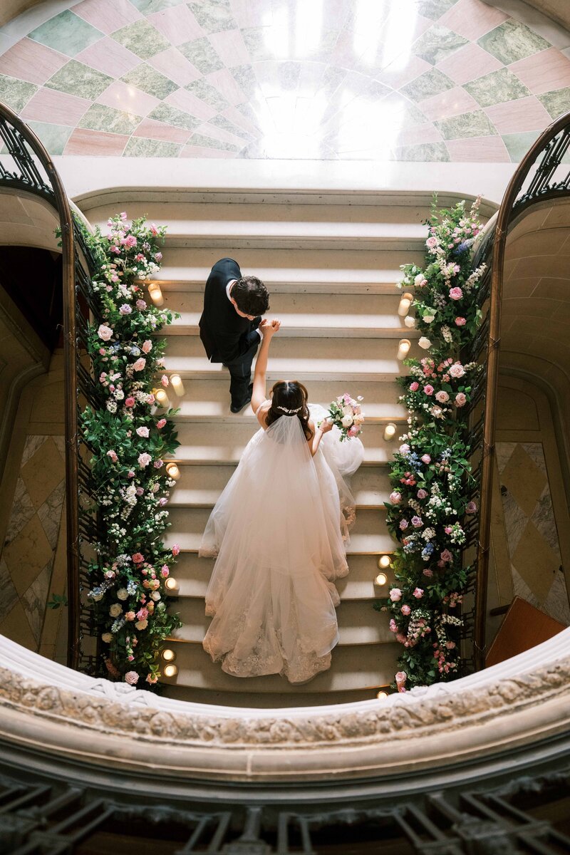 Bride and groom ascending a grand staircase decorated with lush floral arrangements on either side, captured from above.