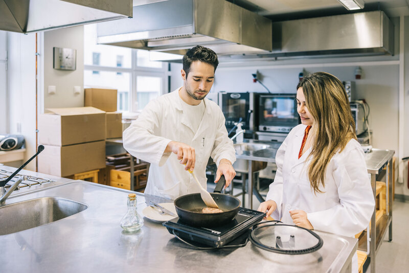 man and woman in a kitchen with lab coats cooking and testing meat 