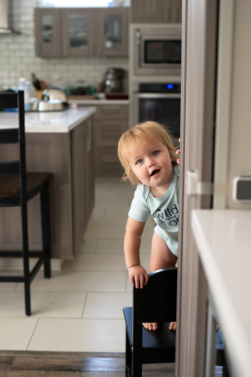 Toddler boy stading on a small chair peeking around the cabinet in the kitchen during an in home baby milestone session