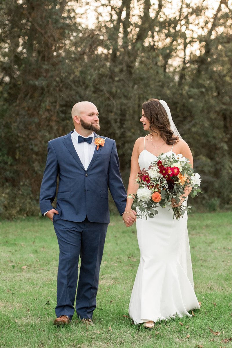 The bride and groom stand in a green field with tall pine trees behind them. The groom has one hand in his pocket and holds the brides hand with the other. He is wearing a navy suit with a white shirt and black bow tie. He has a peach boutonniere pinned to his lapel. The bride and groom look at each other adoringly. The bride is wearing a strapless lace mermaid dress with a fitted bodice and short sweep train.  She is wearing a long sheer veil and is holding a large colorful bouquet. The bouquet has white, red, peach and orange flowers with lush greenery.