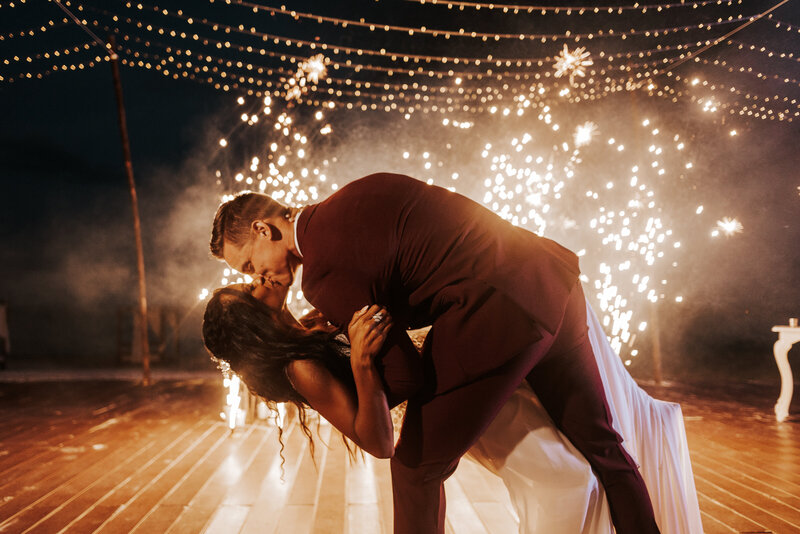 A couple shares a romantic dip and kiss under twinkling lights, with fireworks creating a magical backdrop.