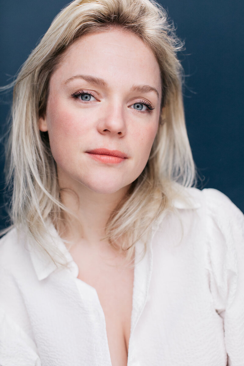 Casting Headshot van musicalactrice met blond haar in witte blouse voor donkerblauwe backdrop.
