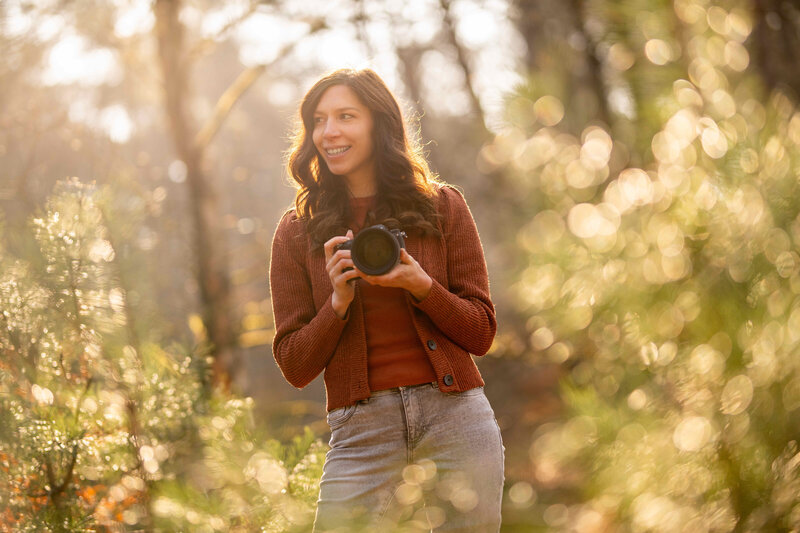 Natascha staat met haar camera in de hand, kijkt opzij in het bos terwijl de zon straalt op de achtergrond