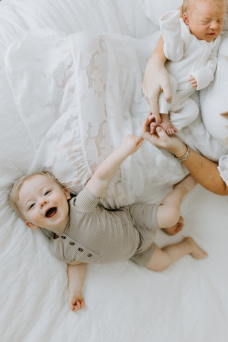 A cheerful toddler lays on the bed with his mother and newborn baby brother with West Lafayette family photographer Vanessa Stoller capturing the photo. 