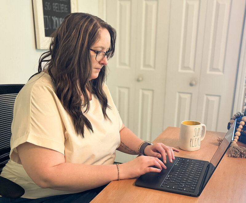 Ali working at her desk with focused expression, typing on black laptop and coffee mug beside her.