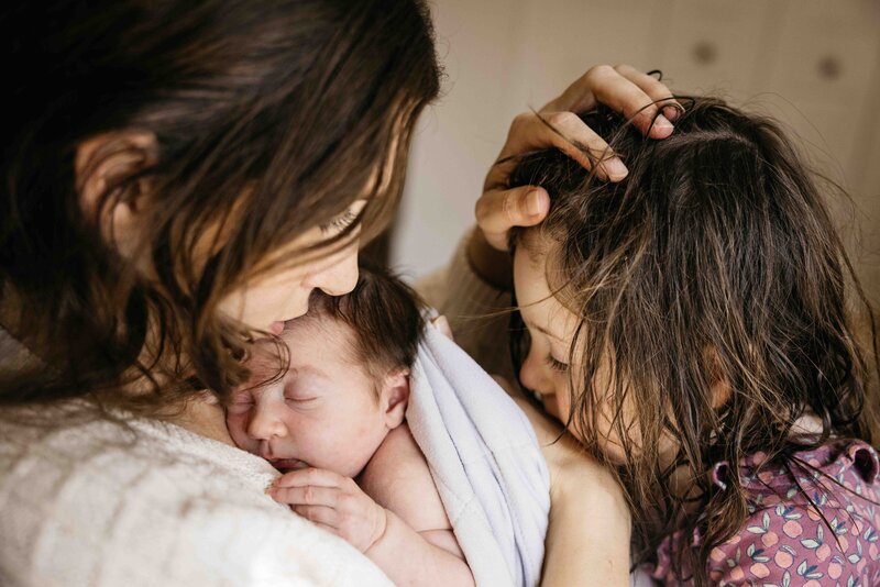 Mom is holding newborn baby and older toddler daughter on her lap. 