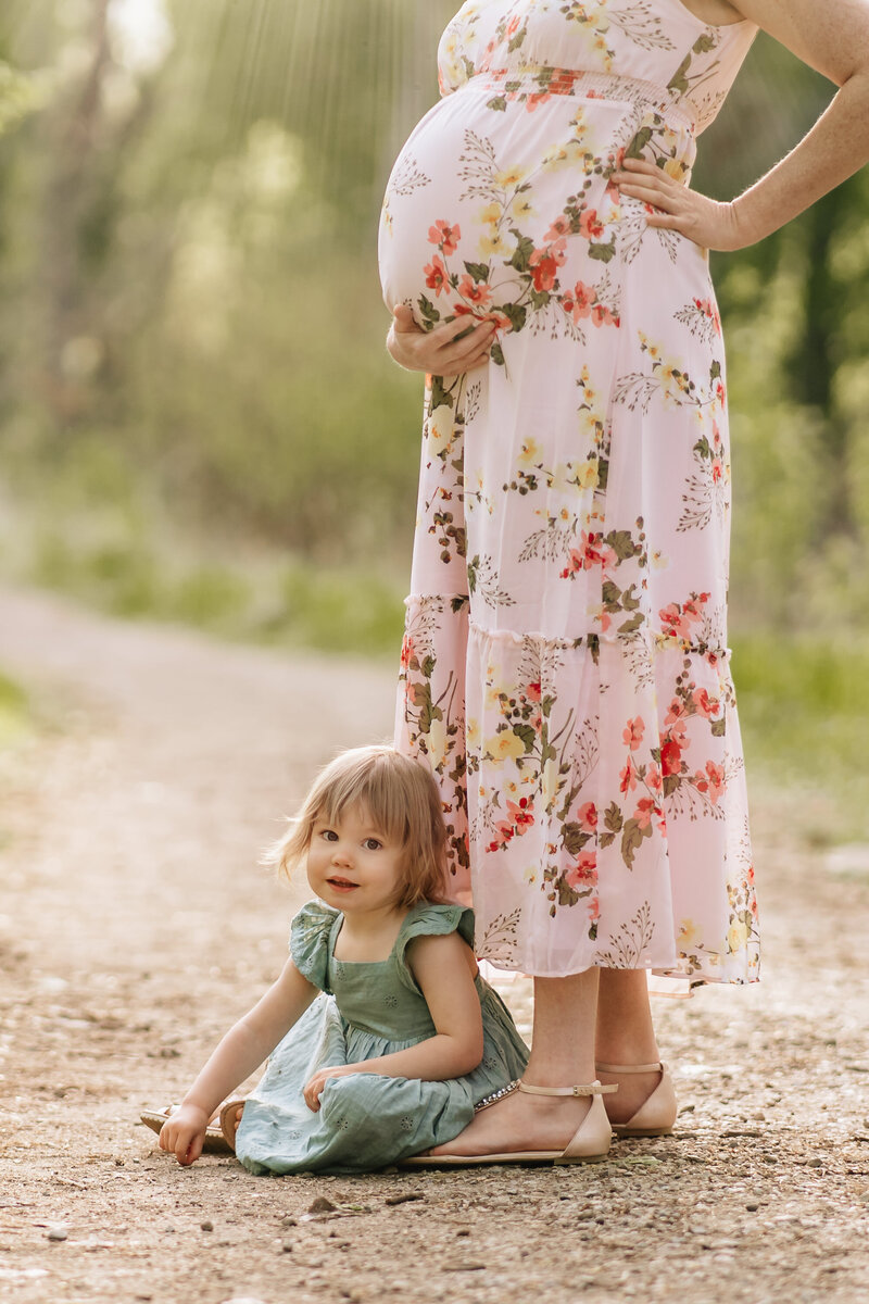 A close-up of a young child in a green dress sitting on the ground next to a pregnant woman in a floral dress, with the focus on the child’s sweet expression and the woman’s baby bump.