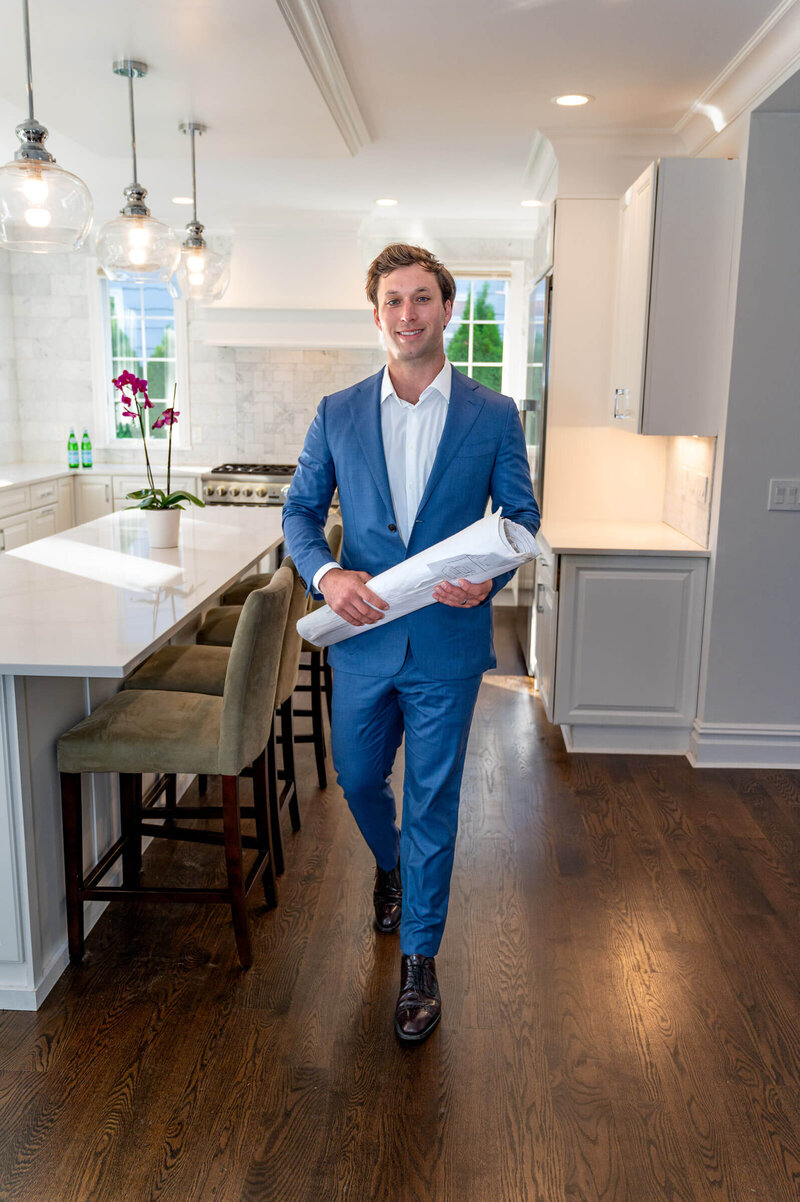 Real estate agent in a newly renovated white kitchen, walking towards the camera, smiling while holding architectural building plans for the house in Greenwich, CT.