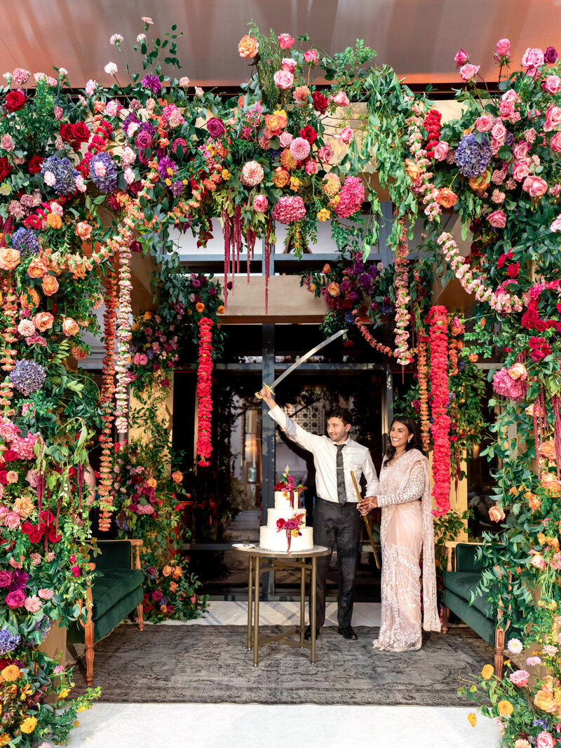 A couple stands under a vibrant floral mandap at a wedding, with the man holding a sword and cutting a white cake on a small table. The woman holds his arm, dressed in an elegant outfit. Colorful flowers adorn the scene, creating a festive atmosphere.