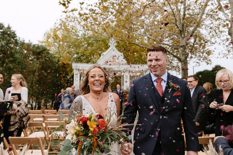 A couple walks hand-in-hand down a wedding aisle, surrounded by guests and colorful flower petals falling from above.