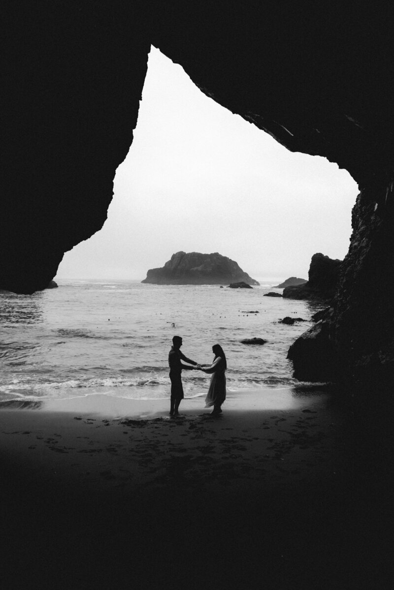 During their Oregon coast elopement, a bride and groom hold hands under the opening of a large sea cave.