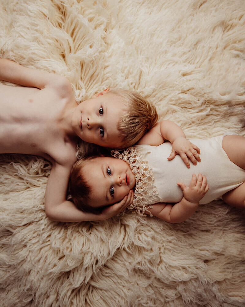 A young boy and his baby sibling lie on a fluffy cream-colored blanket, with the boy protectively holding the baby's head as they both gaze at the camera.