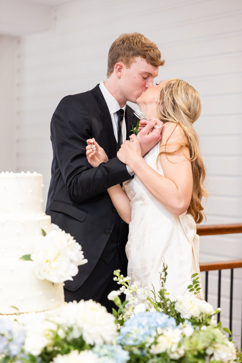 Spring wedding at WinMock at Kinderton, designed by Charming Carolina — bride and groom kiss after cutting their four-tier white wedding cake decorated with pearl accents.