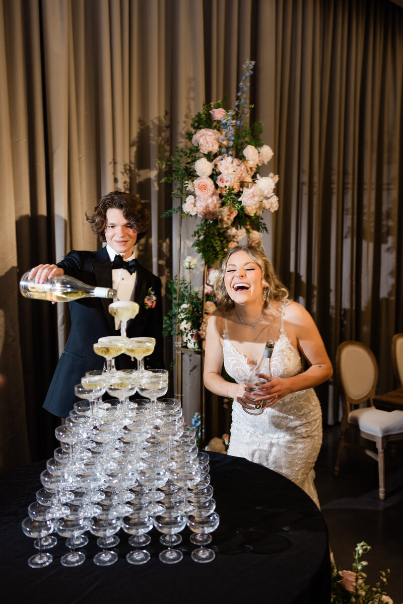 Mini round folding table holding a stacked champagne tower as a bride and groom laugh and pour bubbly during an indoor reception.