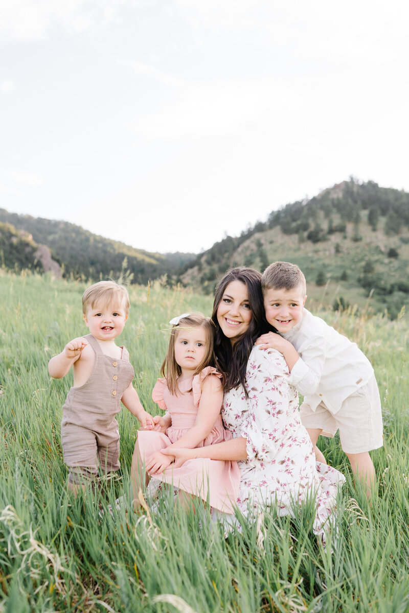 Mom of three snuggling with her children snuggling in tall grass in front of a montain backdrop- photo by Maegan R Photography