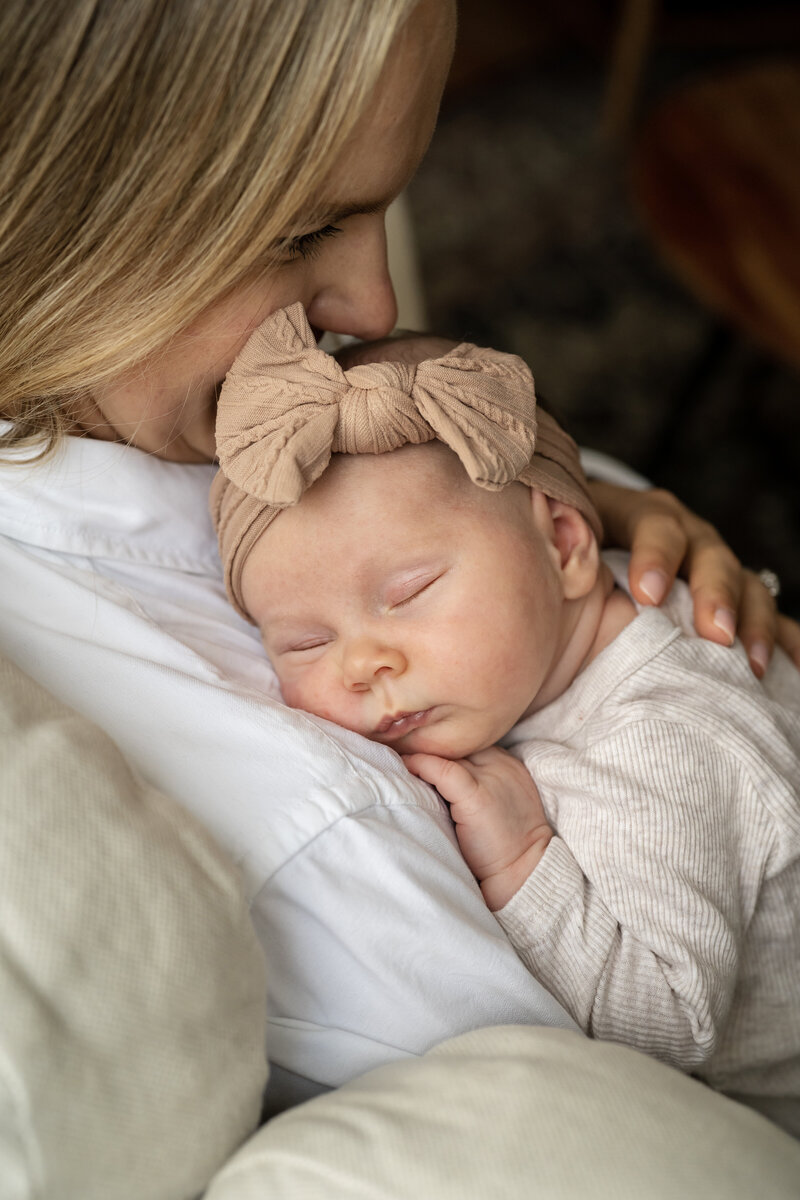 A newborn baby girl sleeps on her mother's shoulder at their home. The baby girl wears a pink headband with a bow.