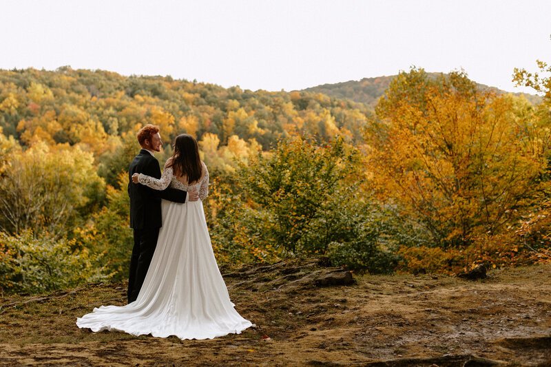 A couple stands together in a serene autumn landscape, surrounded by vibrant trees in shades of orange and green.